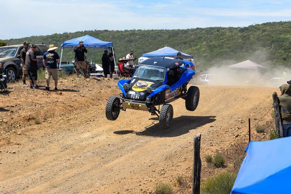 A blue off-road vehicle jumps over a dirt trail during a race.